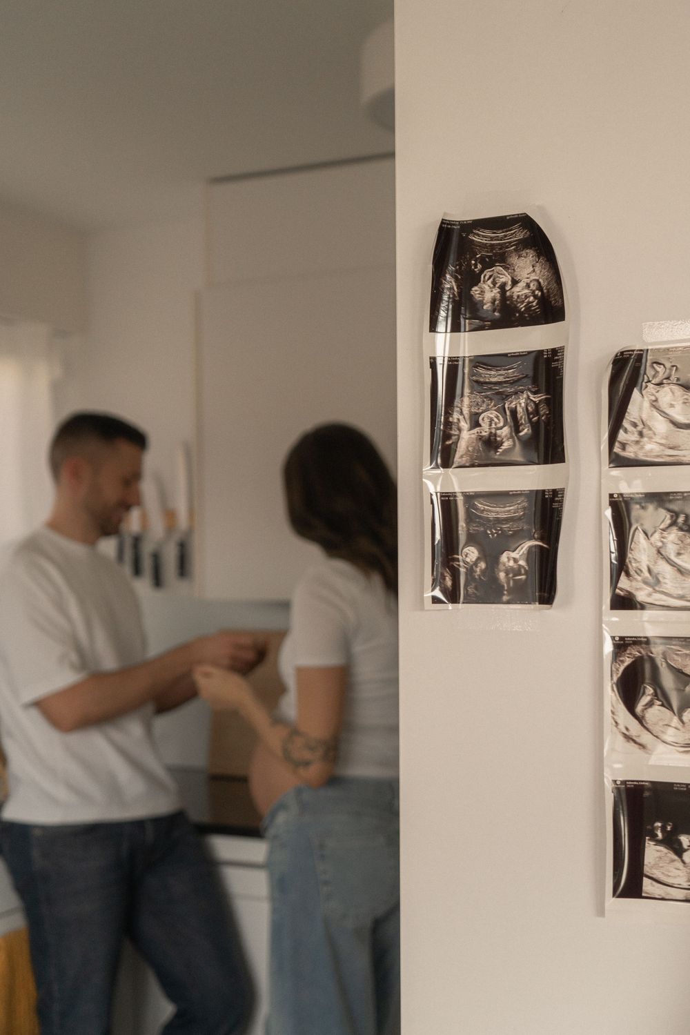 Ultrasound scans taped to a wall with a couple sharing a tender moment in a softly blurred kitchen background.