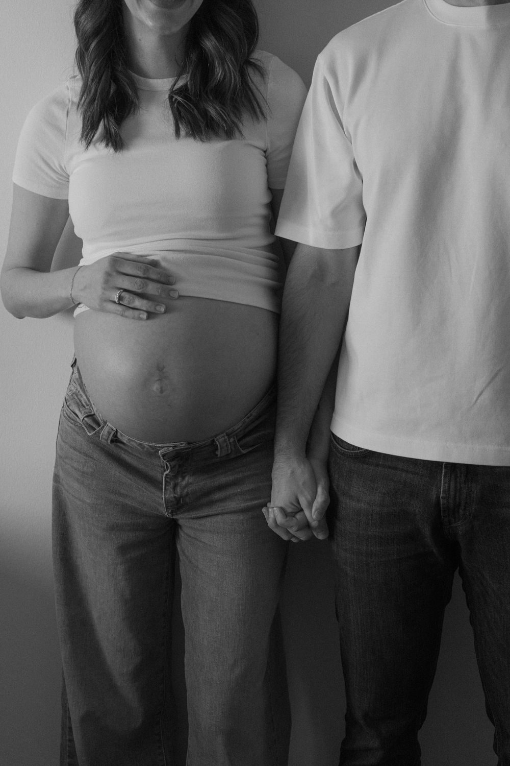 Black and white maternity photo of pregnant woman holding baby bump while partner holds her hand.