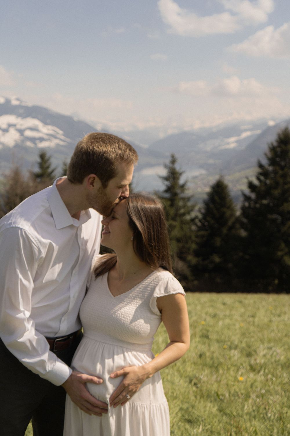 Couple sharing a tender moment on a mountain meadow, man kissing pregnant woman's forehead while holding her baby bump.