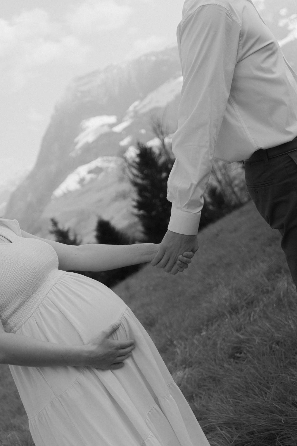 Black and white photo of pregnant woman's baby bump as couple holds hands on a scenic mountain hillside.