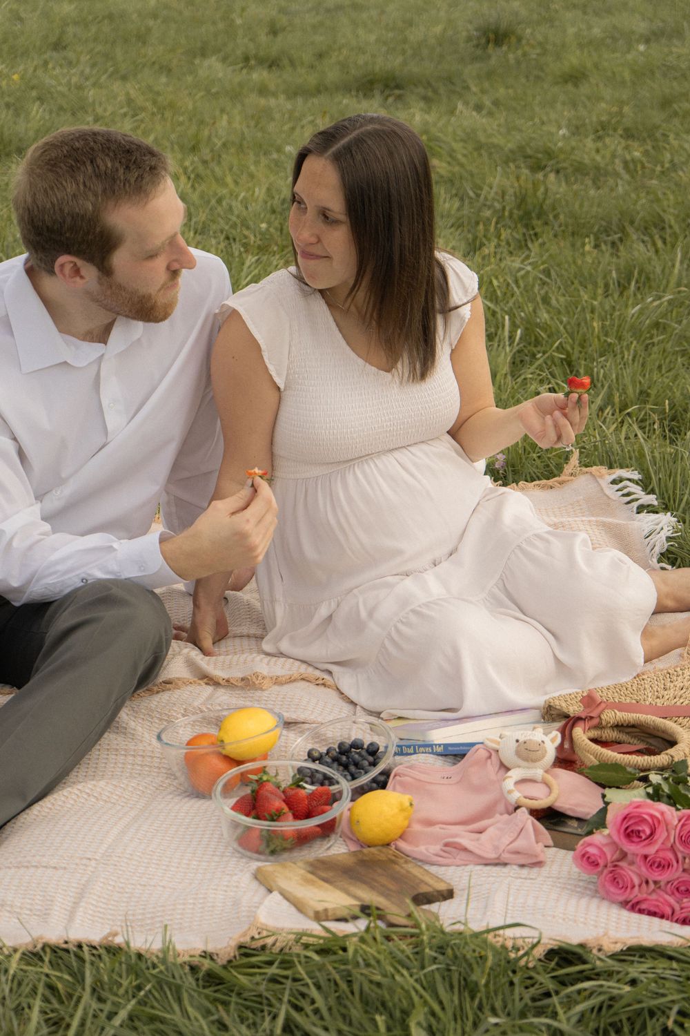 Pregnant woman in white dress enjoying a romantic outdoor picnic with her partner, eating strawberries on a blanket in a green field.