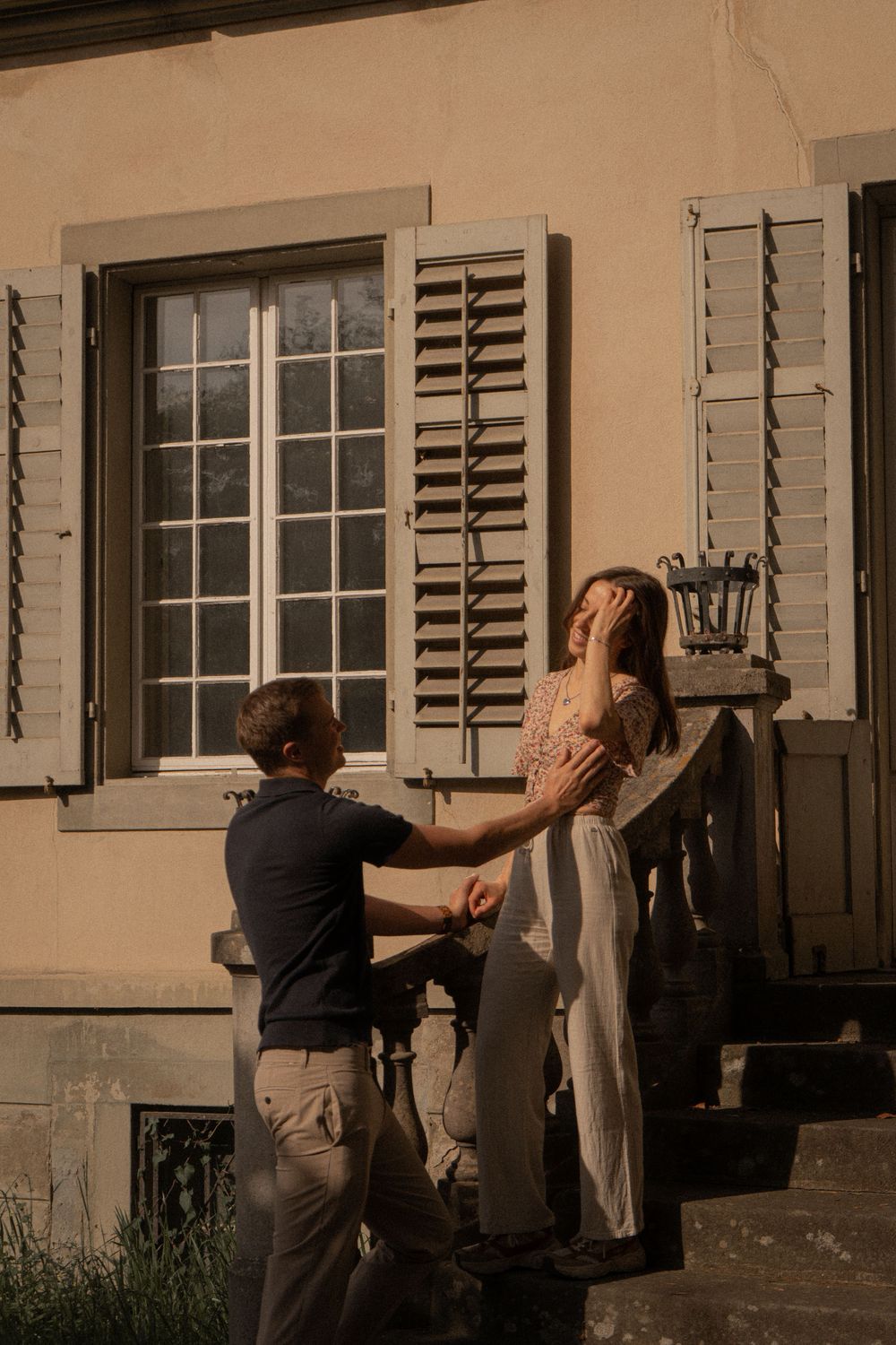 Couple sharing a romantic moment on sunlit steps outside a classic European building with shuttered windows.