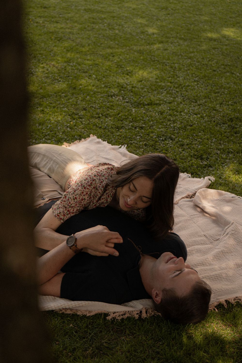 Couple relaxing on a blanket in green grass park, woman smiling down at man lying on her lap in golden sunlight.