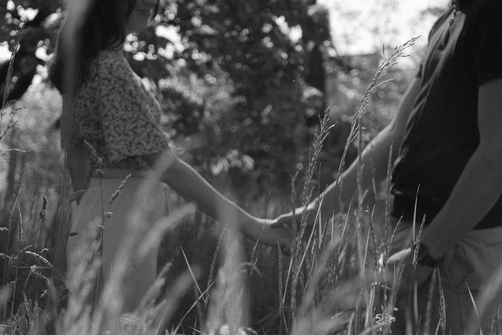 Black and white photo of a couple holding hands while walking through tall grass in a sunlit field.