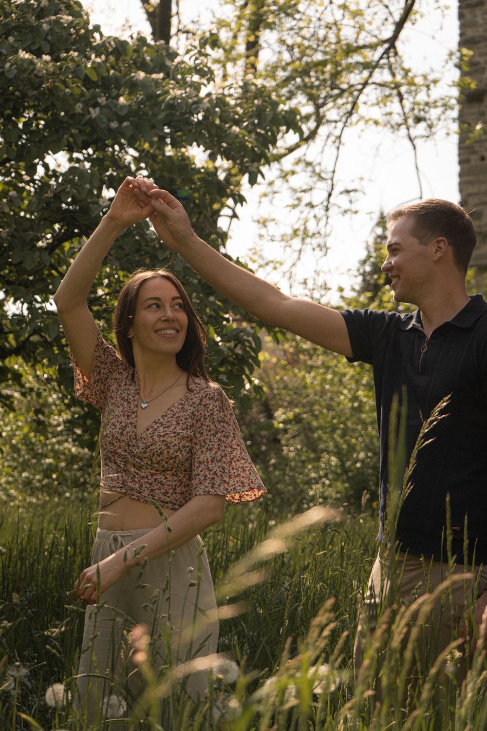 A couple dancing joyfully in a sunlit green meadow, the woman in a floral crop top spinning under her partner's raised hand.