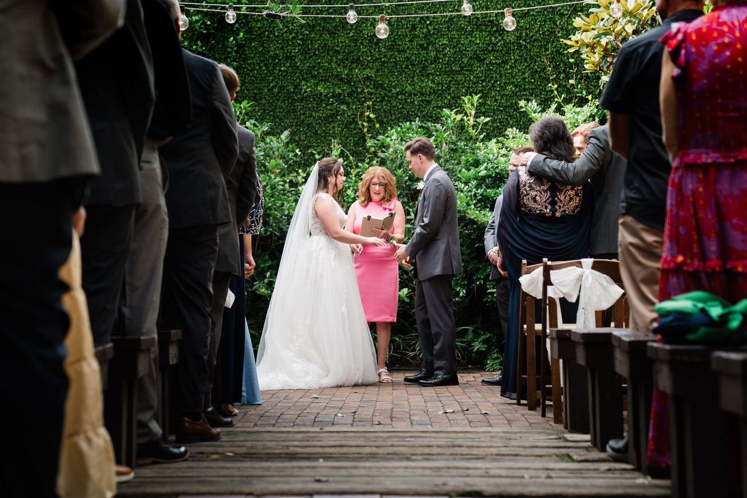 String lights illuminate an outdoor wedding ceremony aisle lined with guests in a garden setting.