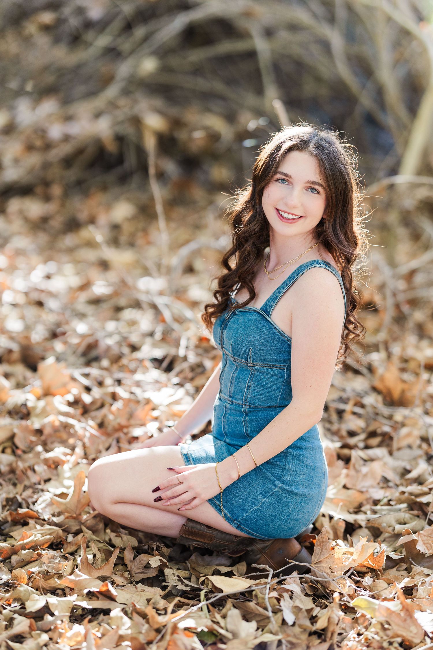 A person in a denim dress poses on autumn leaves in a natural outdoor setting during a photo session.