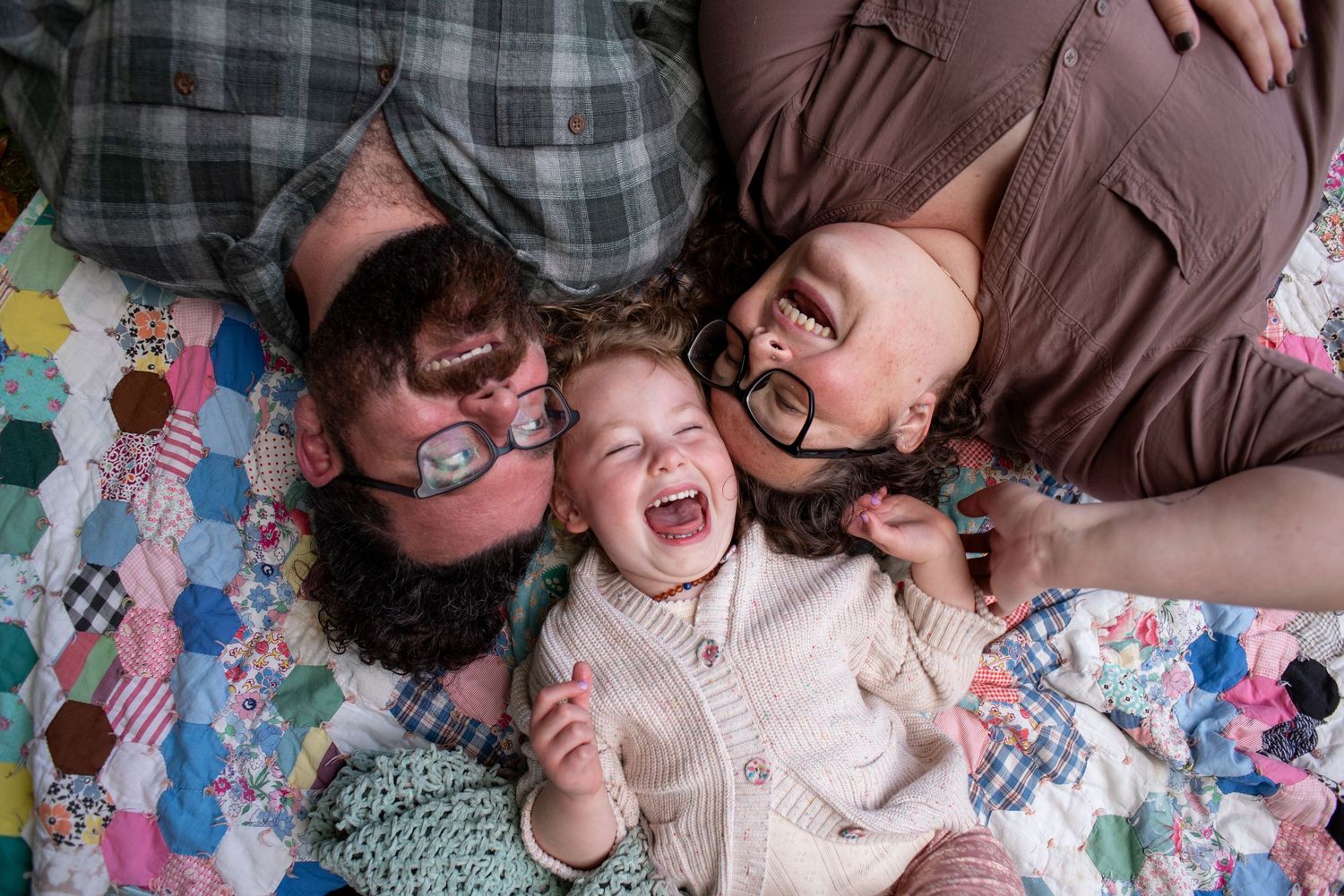 A candid family photograph by Aslyn O Photo of parents and a child laying on a quilt blanket, laughing together in Michigan
