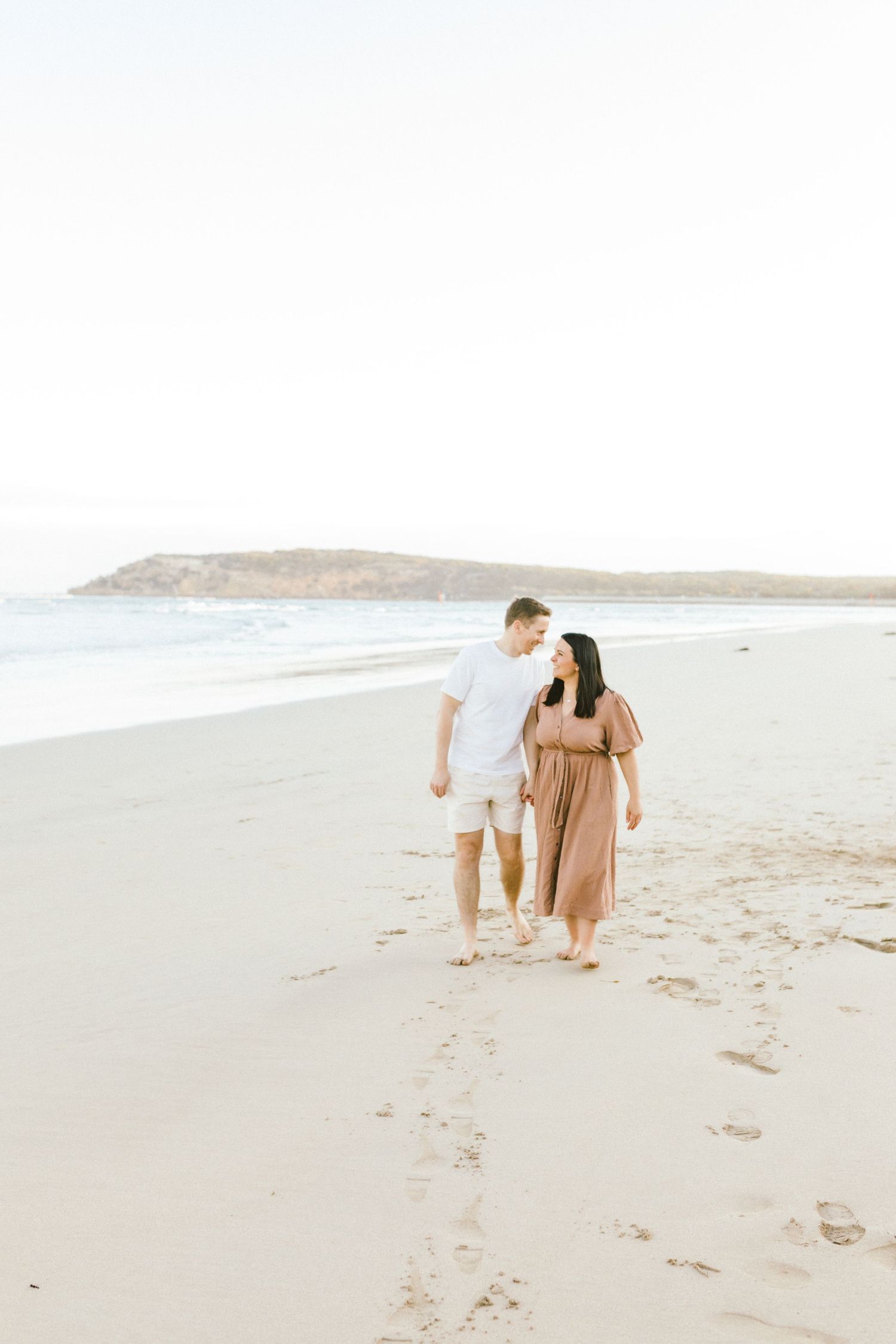 A couple walks along a serene beach shoreline during sunset in neutral-toned outfits.