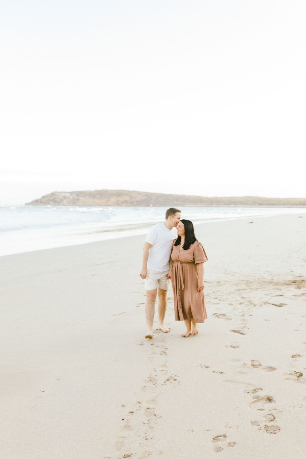 A couple walking together on a serene beach with mountains in the distance during golden hour.