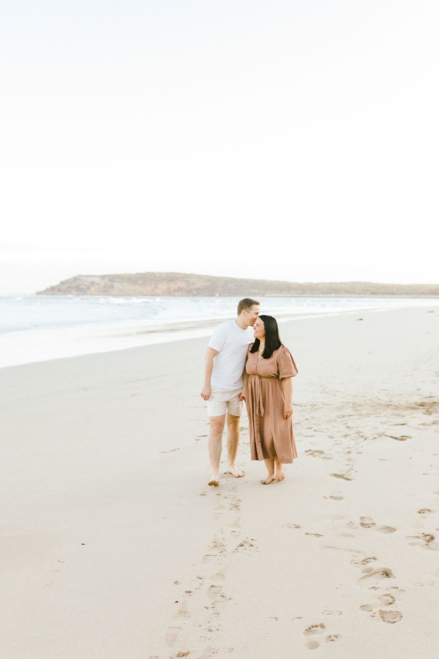 A couple walking together on a serene beach with mountains in the distance during golden hour.