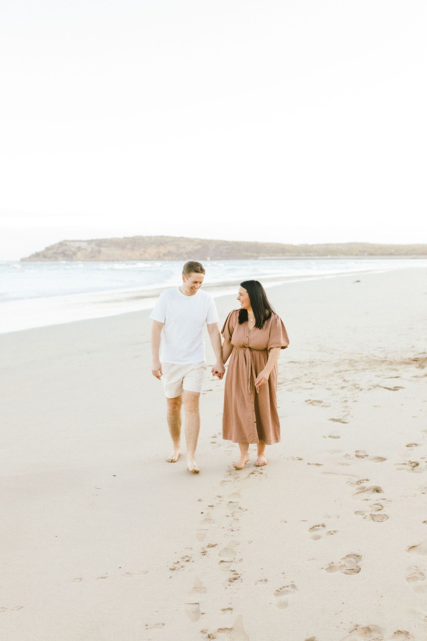 Three people in neutral-toned clothing walk together along a serene sandy beach on an overcast day.