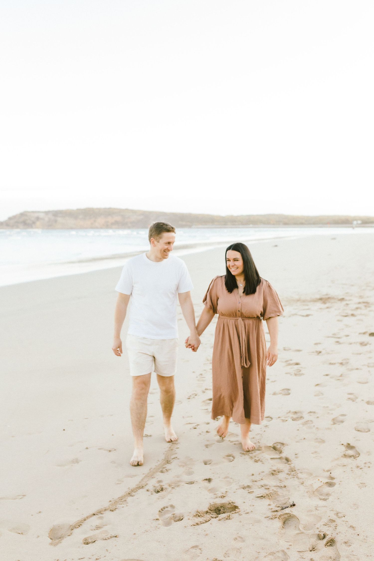 A couple walks together along a sandy beach during sunset, leaving footprints in their wake.