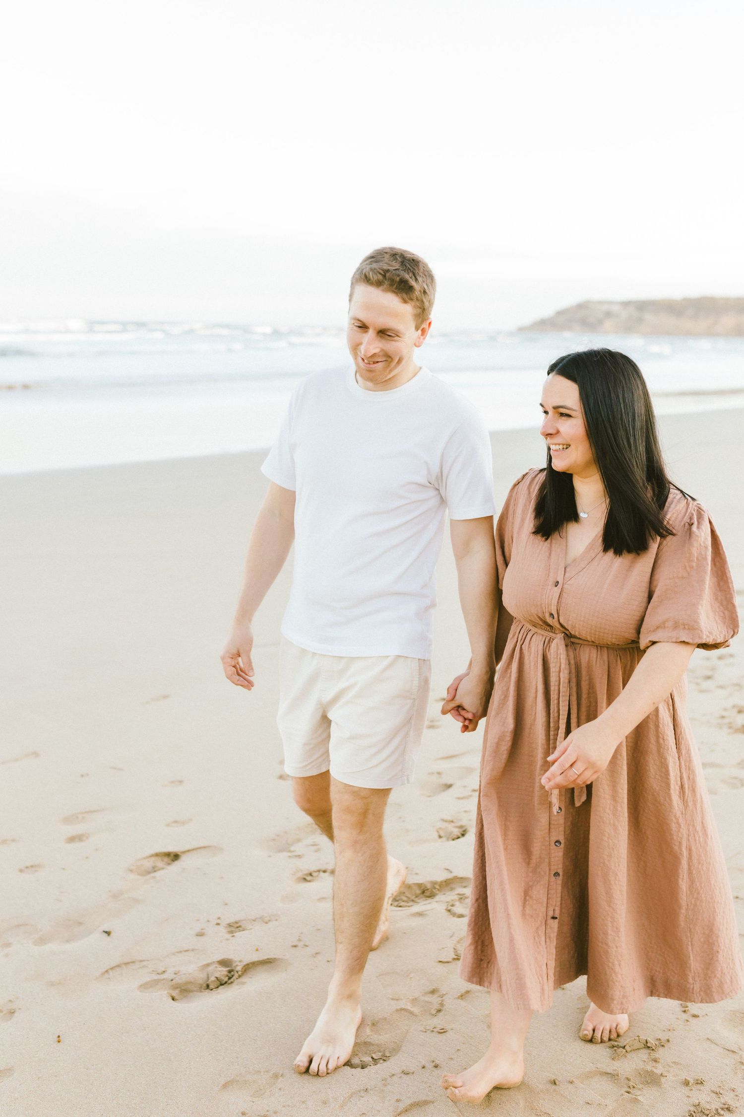 Couple walking barefoot together on sandy beach at sunset in romantic coastal photoshoot.