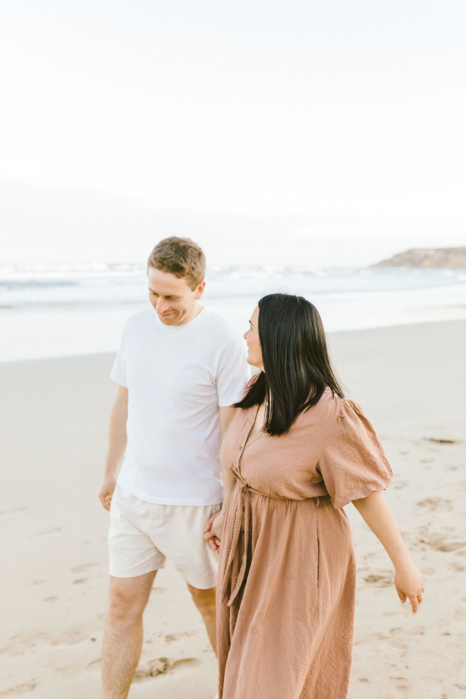 A couple walks together on a sandy beach at sunset, with the person in white shorts and the other in a flowing peach dress.