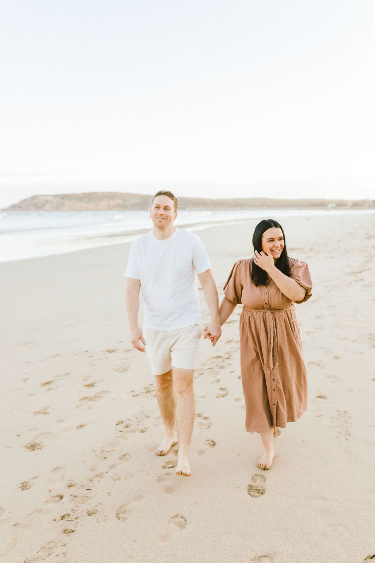 A couple walks together on a sandy beach wearing light colored clothing in a romantic seaside setting.