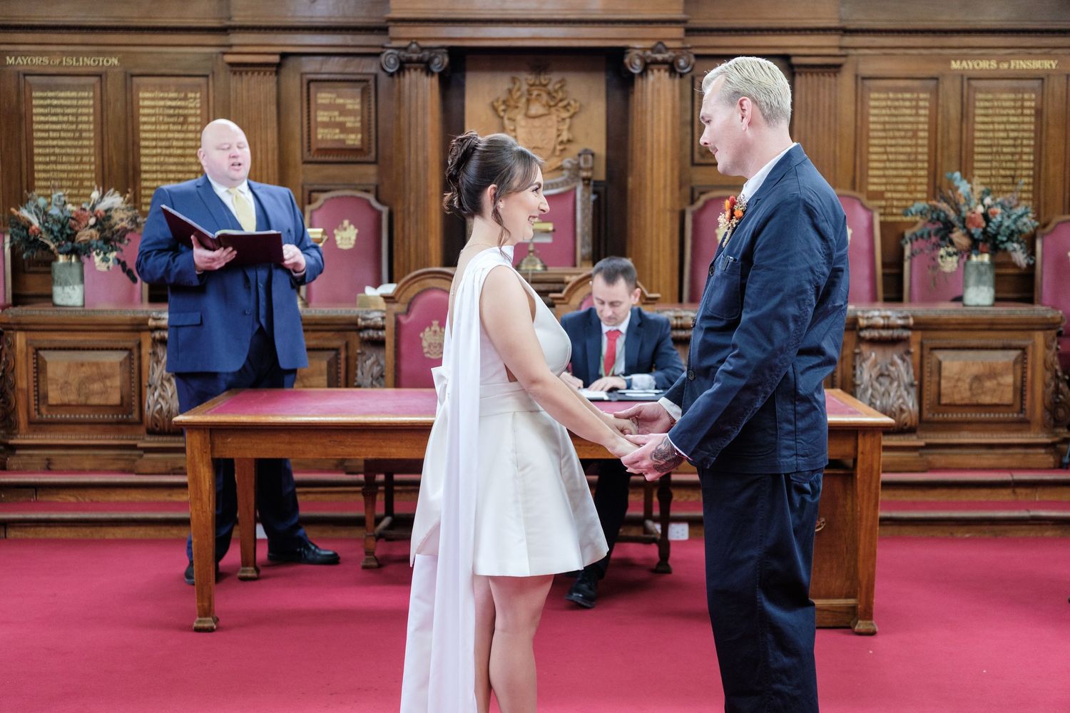 Wedding ceremony takes place in an ornate wood-paneled room with red carpet as couple exchanges rings at the altar.