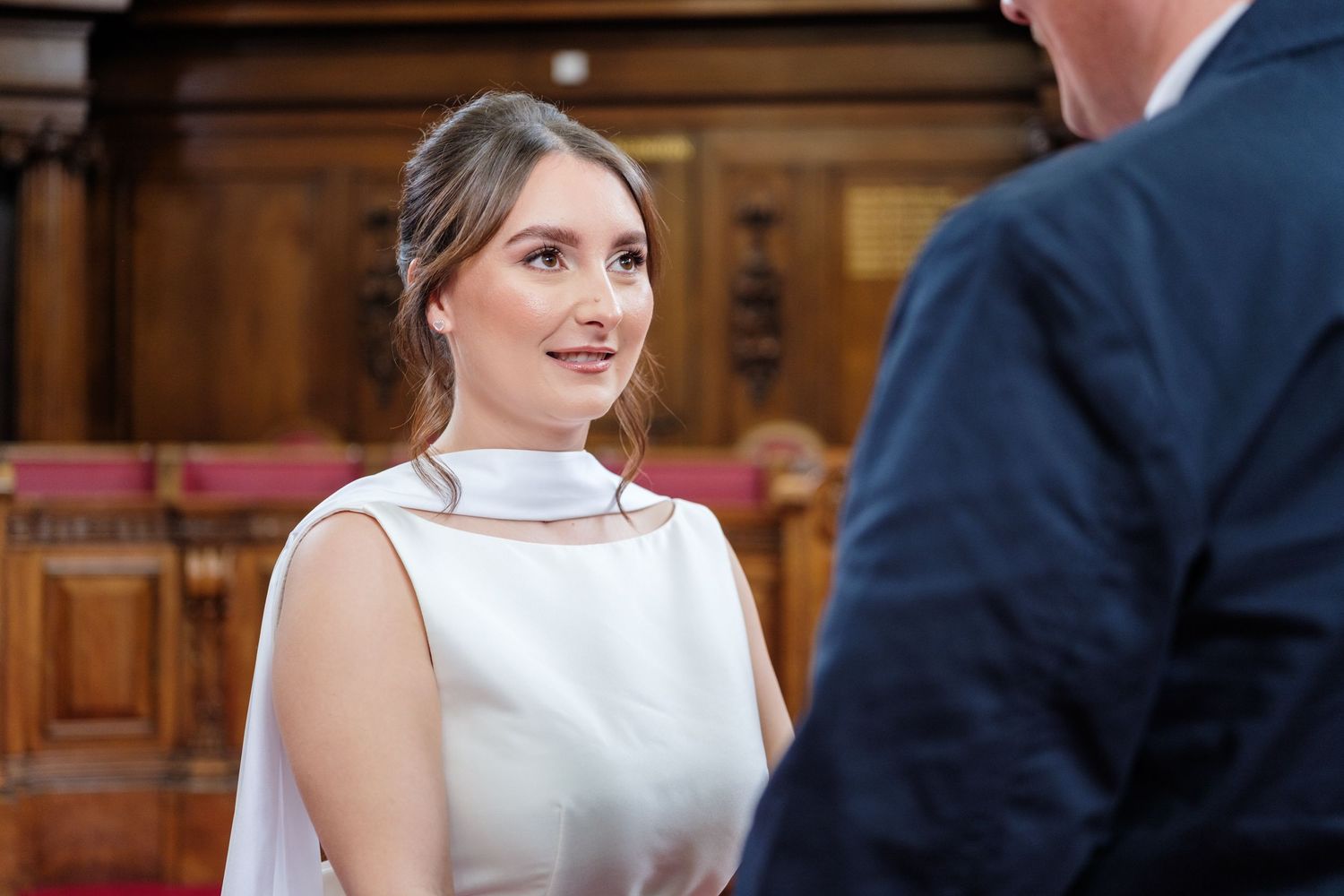 A person in a white sleeveless dress smiles while looking over their shoulder in an elegant indoor setting.