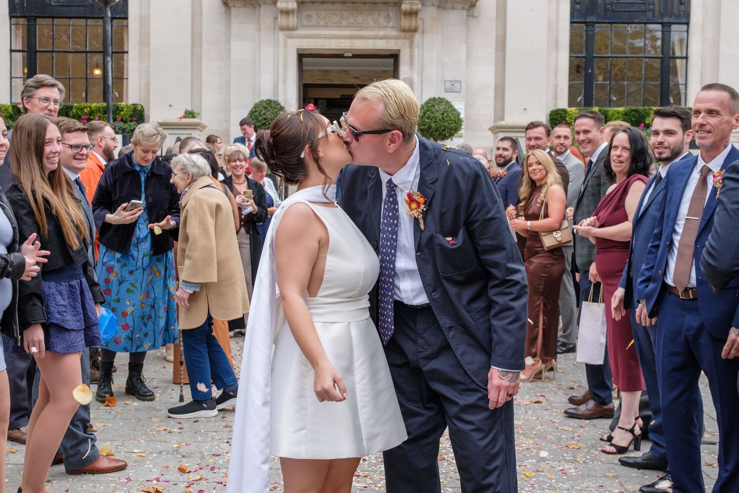 Wedding guests gather outside a stone building throwing confetti at the happy couple during an autumn celebration.