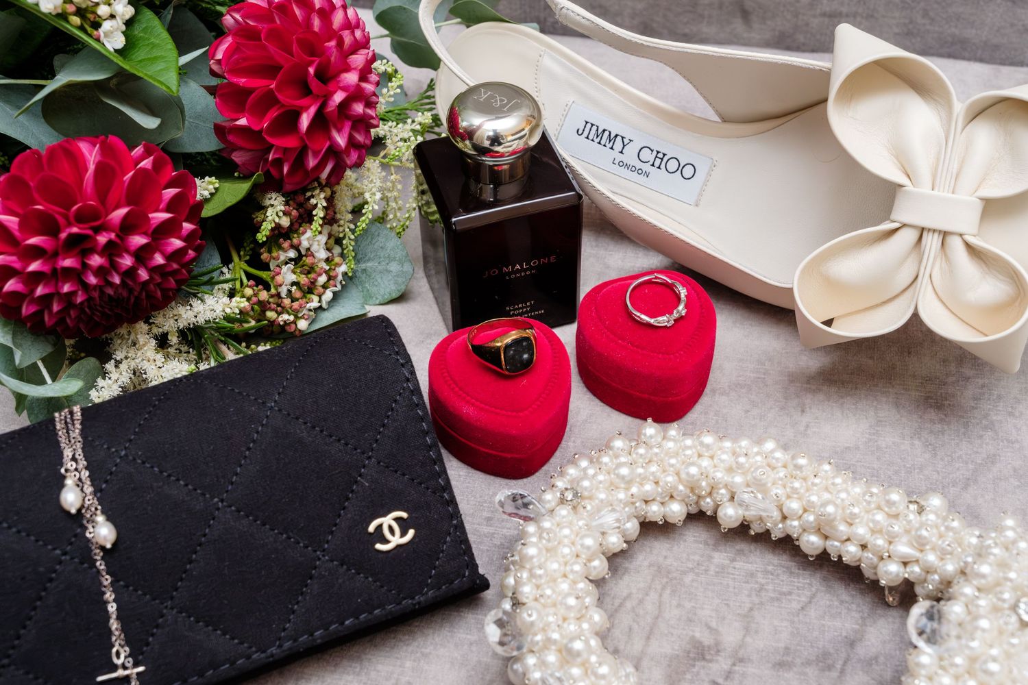 Elegant wedding accessories including red heart ring boxes, black designer purse, pearl necklace and red flowers on grey surface.