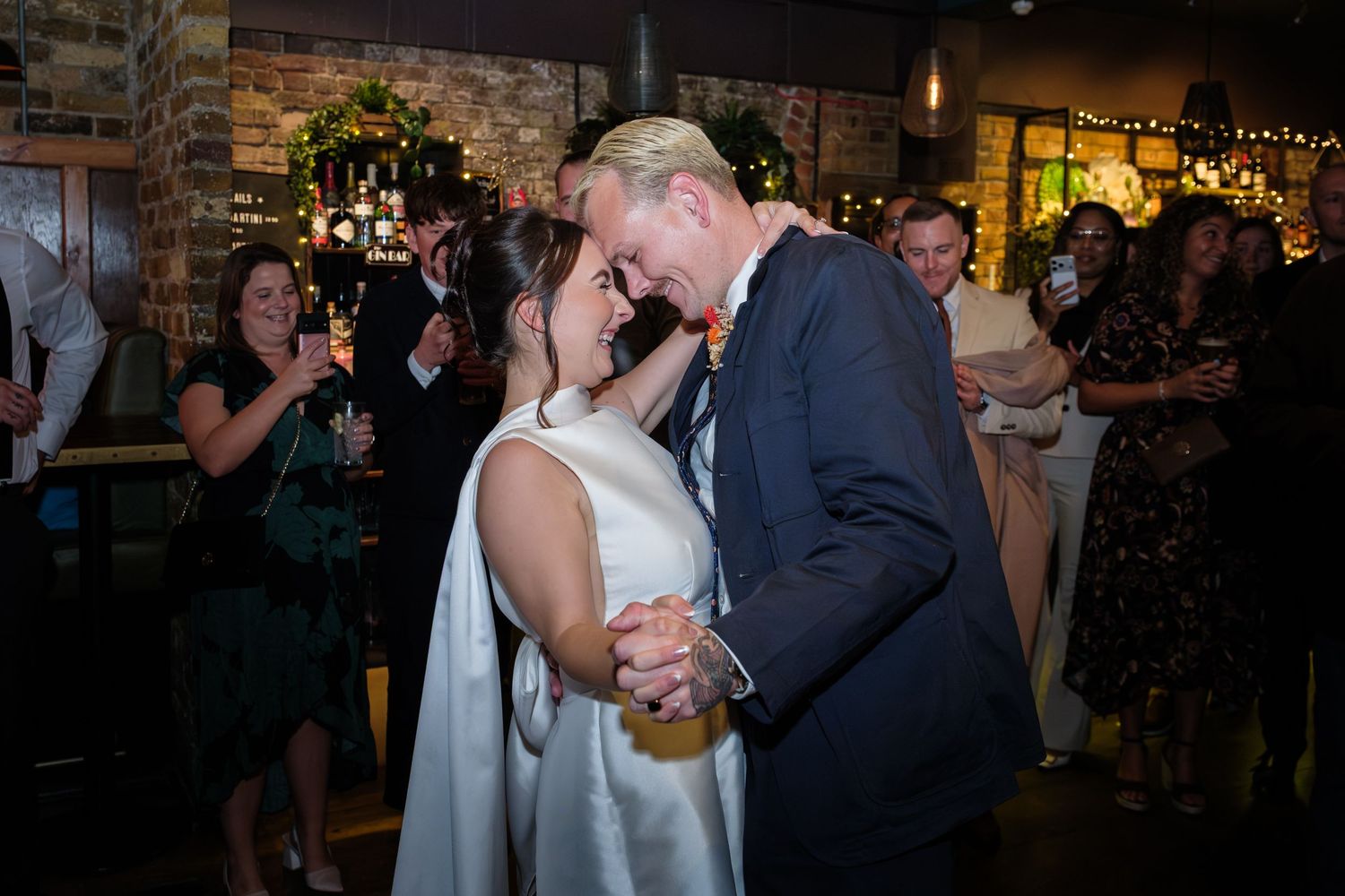 A couple shares a romantic dance at a dimly lit wedding reception with stone walls and decorative lighting.