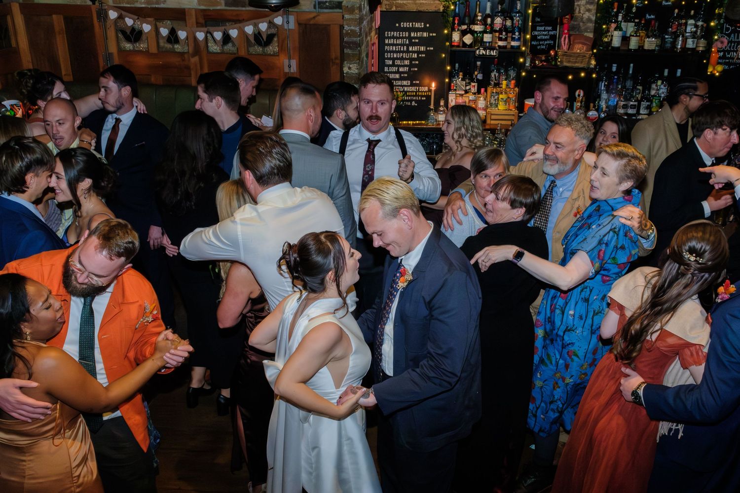 Couples dancing and embracing on a dimly lit dance floor during a festive celebration with string lights overhead.