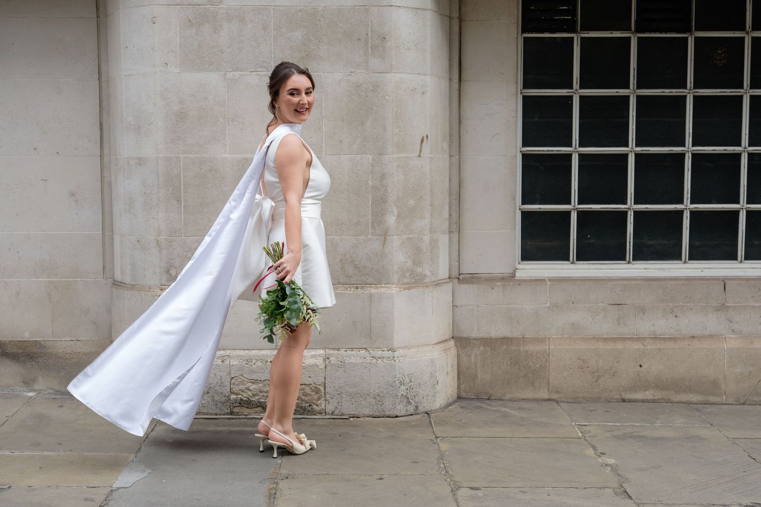 A bride in a flowing white wedding dress and veil walks along a stone building exterior with a bouquet.