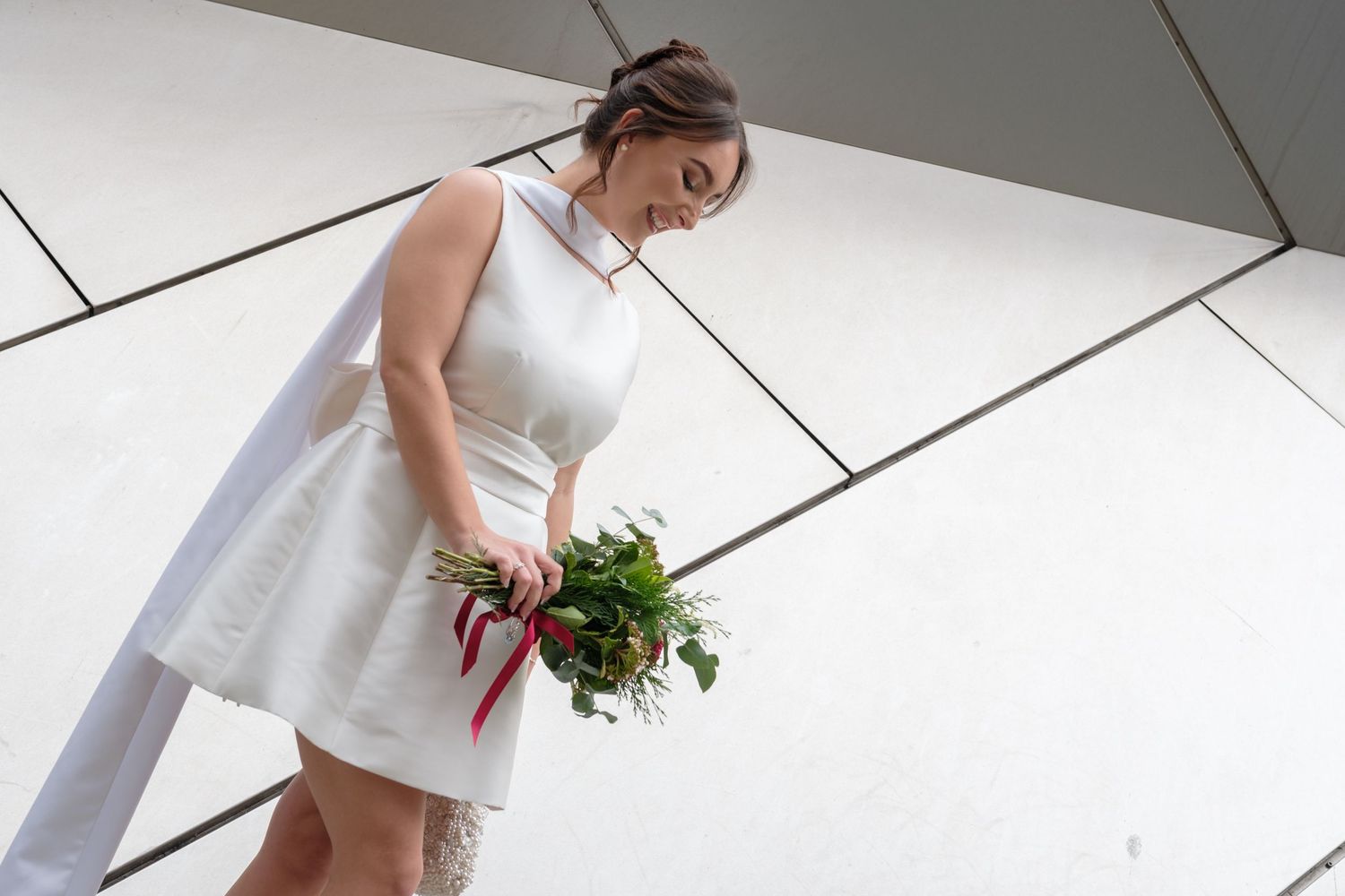 A bride in a white wedding dress holds a bouquet of flowers while walking down a modern hallway.