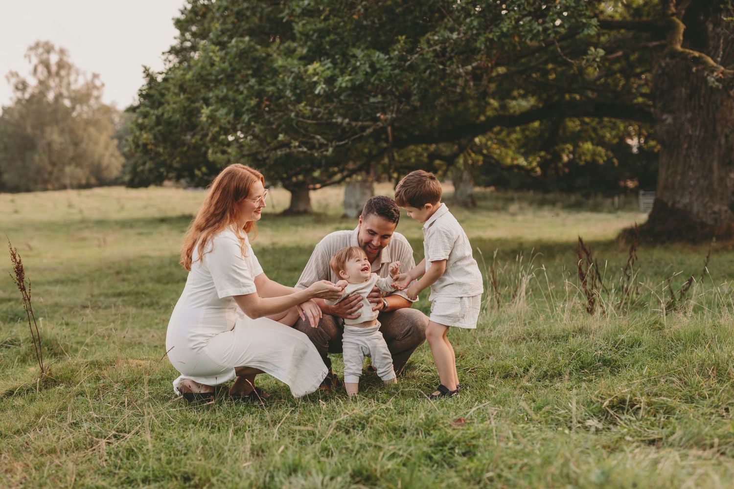 A family in white outfits sits together on grass during a golden hour outdoor photo session.