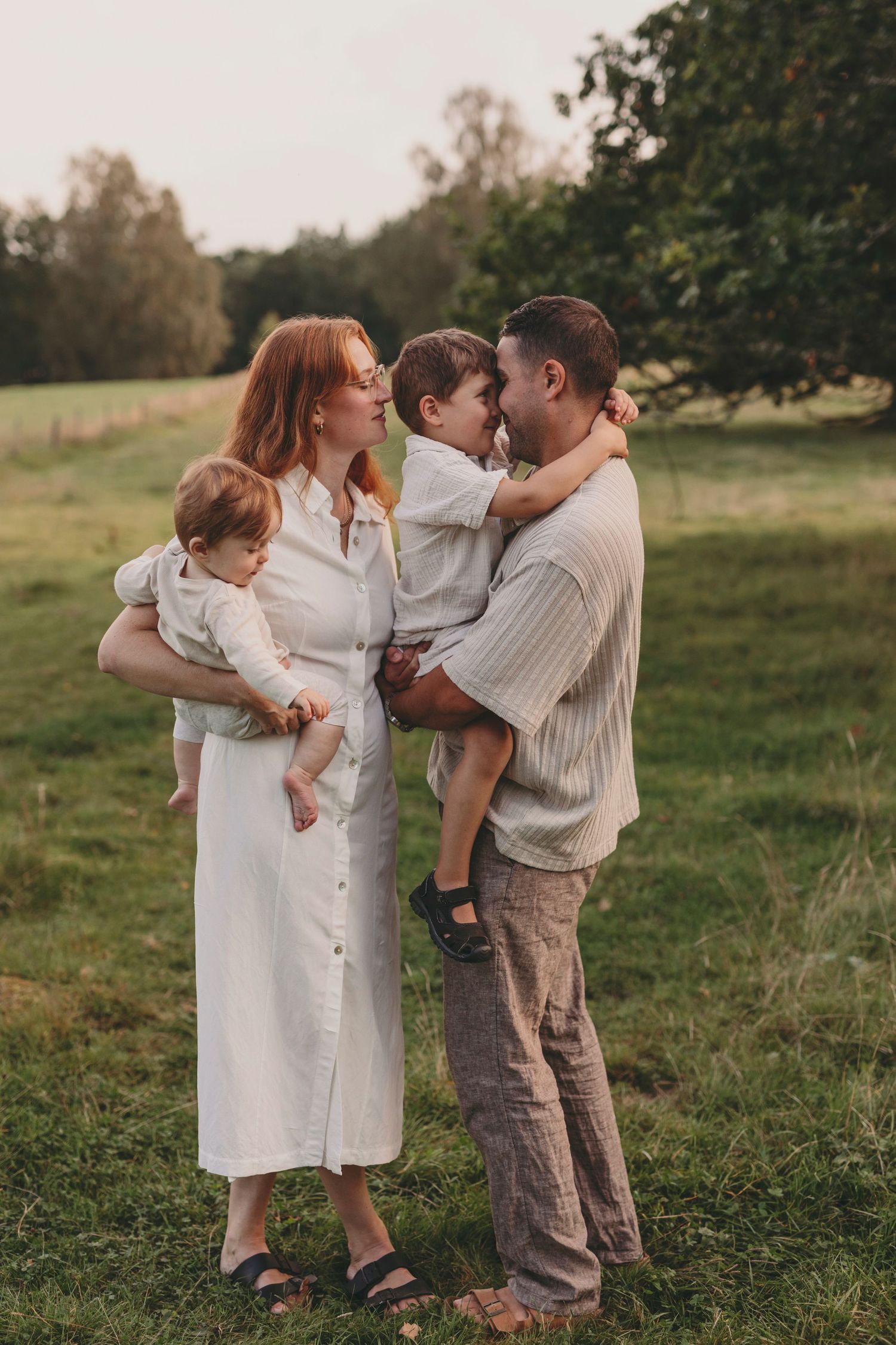 A family shares tender moments together in a grassy field during sunset, wearing casual white and beige clothing.