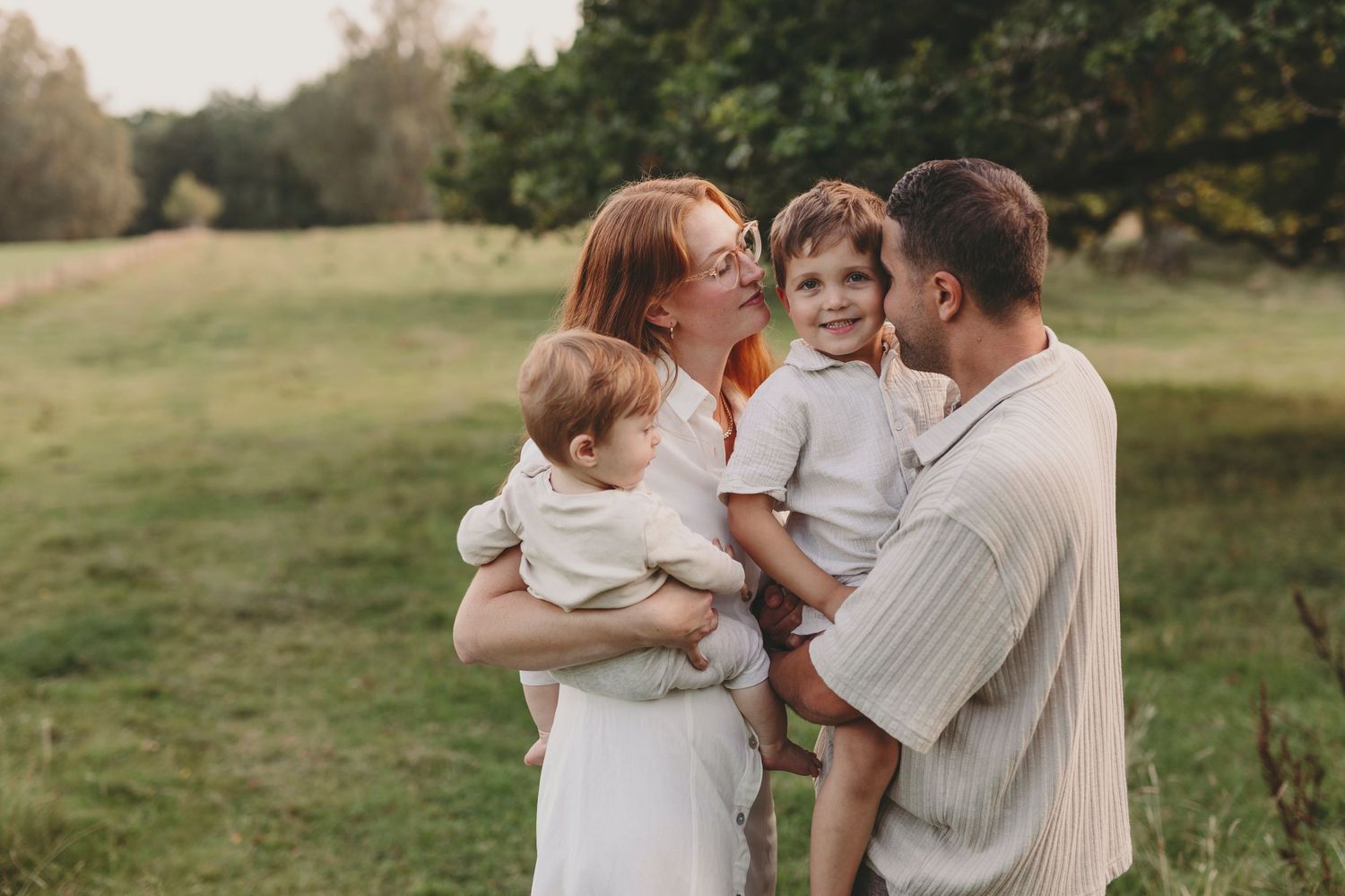 A loving family embraces outdoors in a grassy field during golden hour while wearing neutral colored clothing.