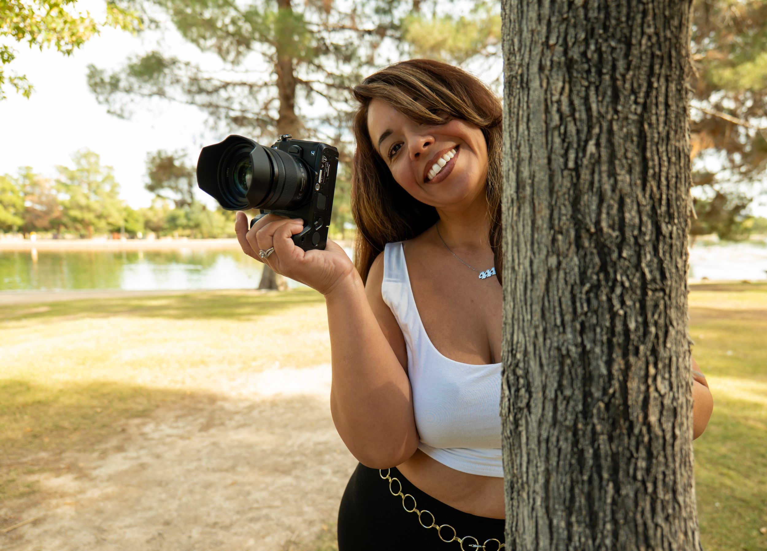 Smiling woman holding a DSLR camera while peeking around a tree in a sunny park.