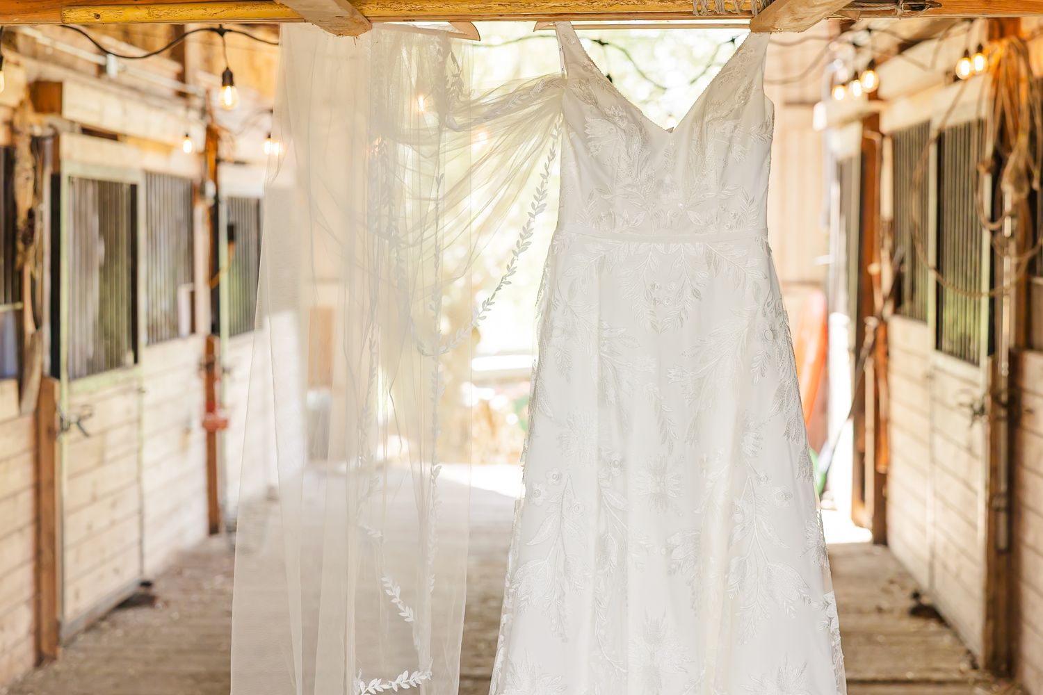 A delicate white wedding dress hangs in a rustic barn hallway with soft natural lighting streaming through.