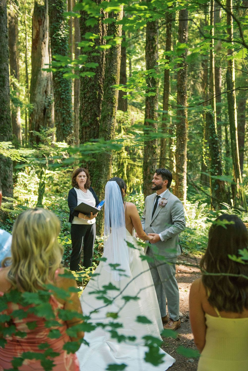 A wedding ceremony takes place on a forest trail surrounded by tall trees and dappled sunlight.