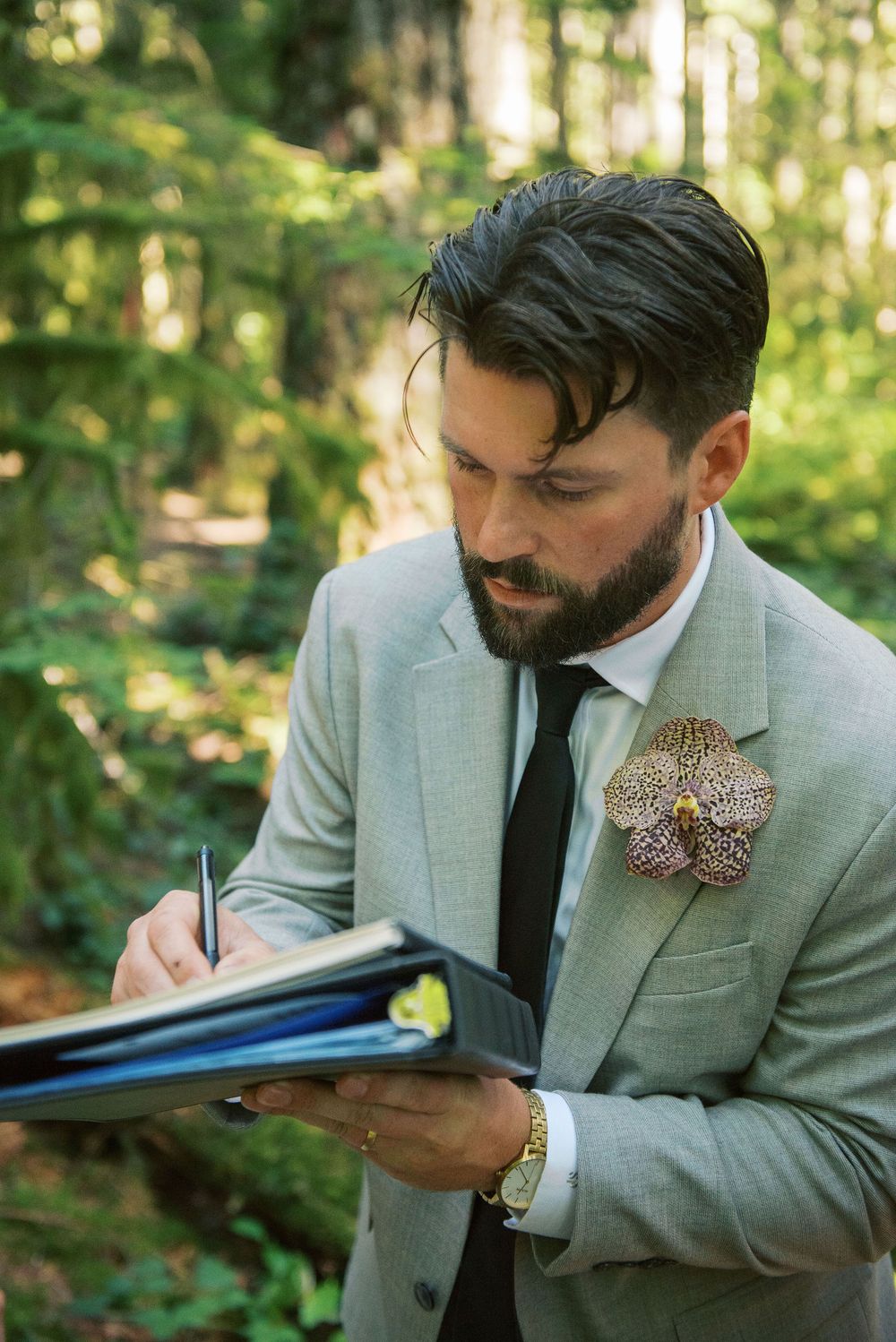A person in a light gray suit with a dark tie signs documents while wearing an orchid boutonniere in an outdoor forest setting.