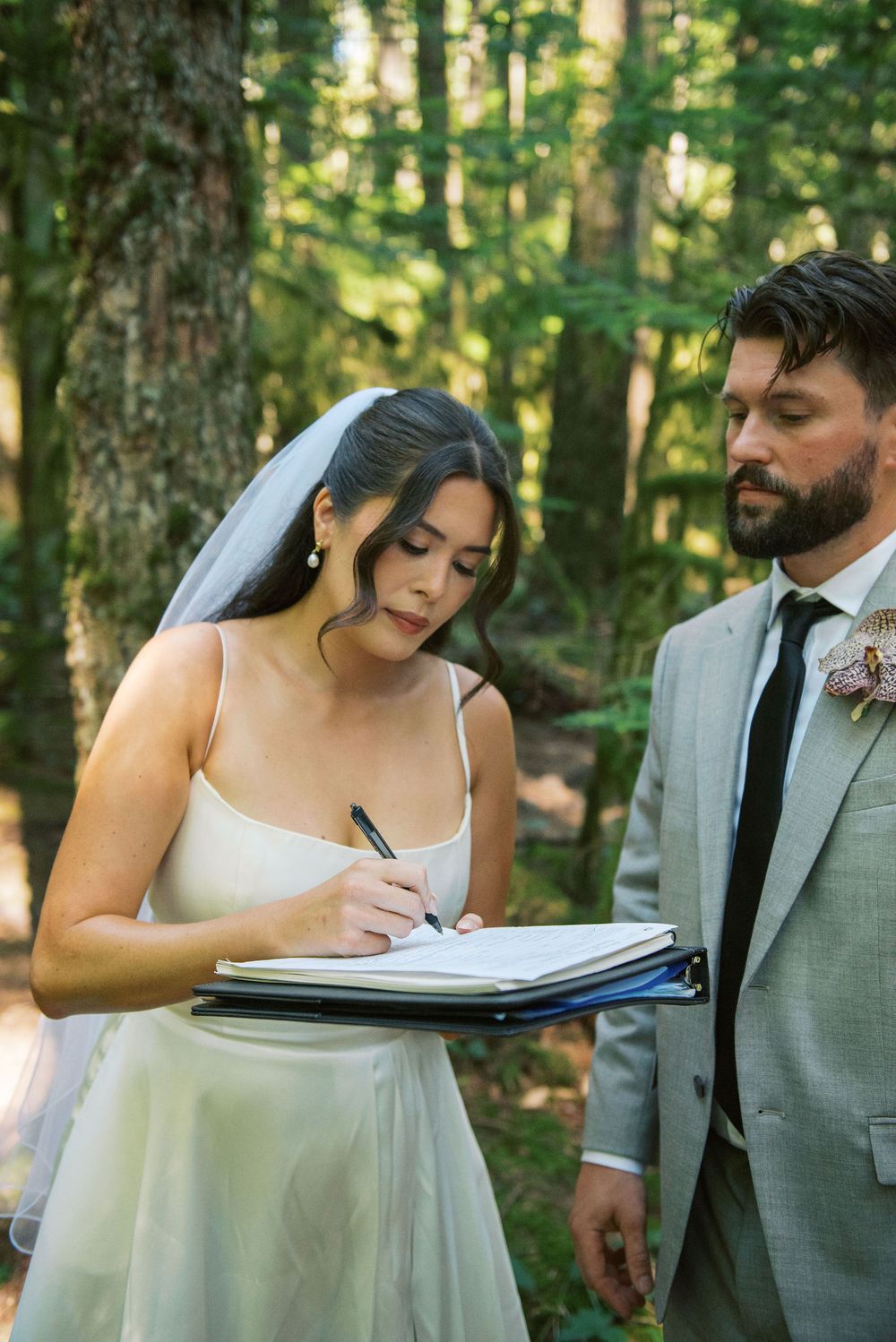 A bride in a white dress signs paperwork during an outdoor forest wedding ceremony.