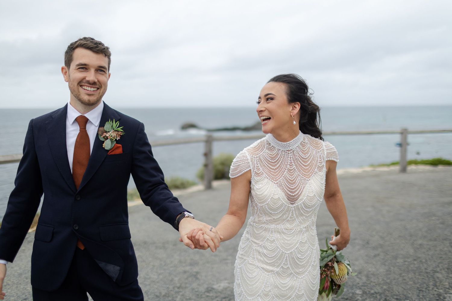 A happy couple holds hands while walking along a coastal path on their wedding day in formal attire.