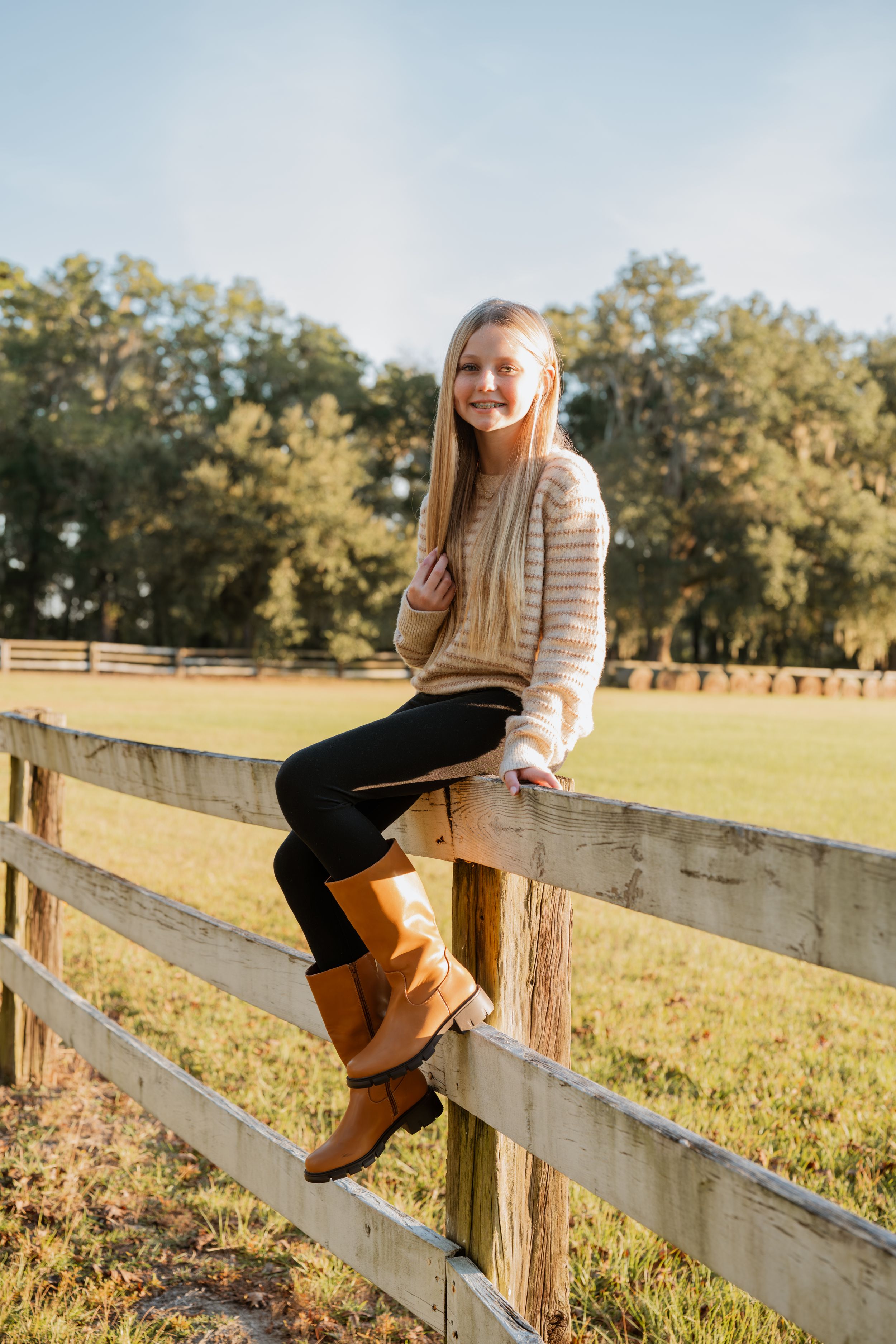 Child in striped sweater sits on wooden fence in sunny field with autumn trees.