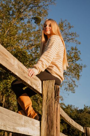 Person in striped sweater perches on rustic wooden fence against blue autumn sky.