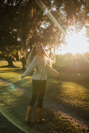 A person in a striped sweater twirls playfully in a sunlit park during golden hour.