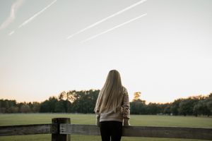 Person in white sweater standing against bright sky at sunset in open field.