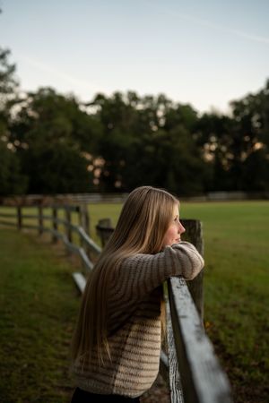 Someone in a knit sweater leans on wooden fence overlooking grassy field at dusk.