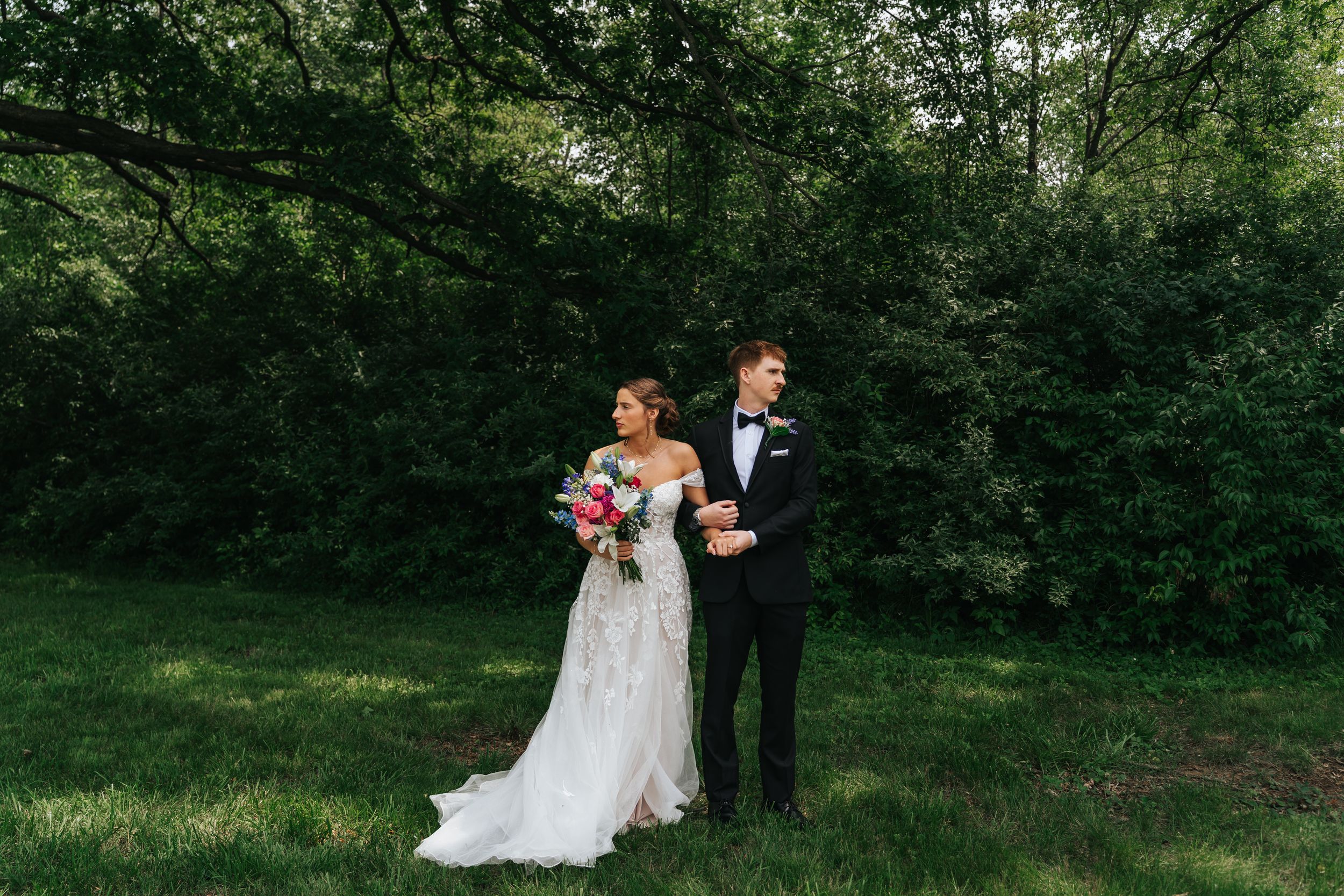 A couple in wedding attire stands together on a lush green lawn surrounded by trees.