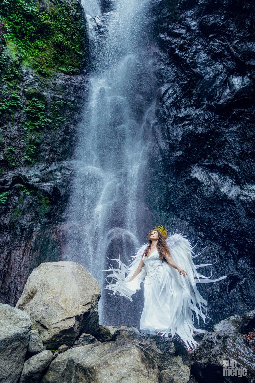 Ethereal figure in flowing white dress poses dramatically against a cascading waterfall in a lush forest setting.