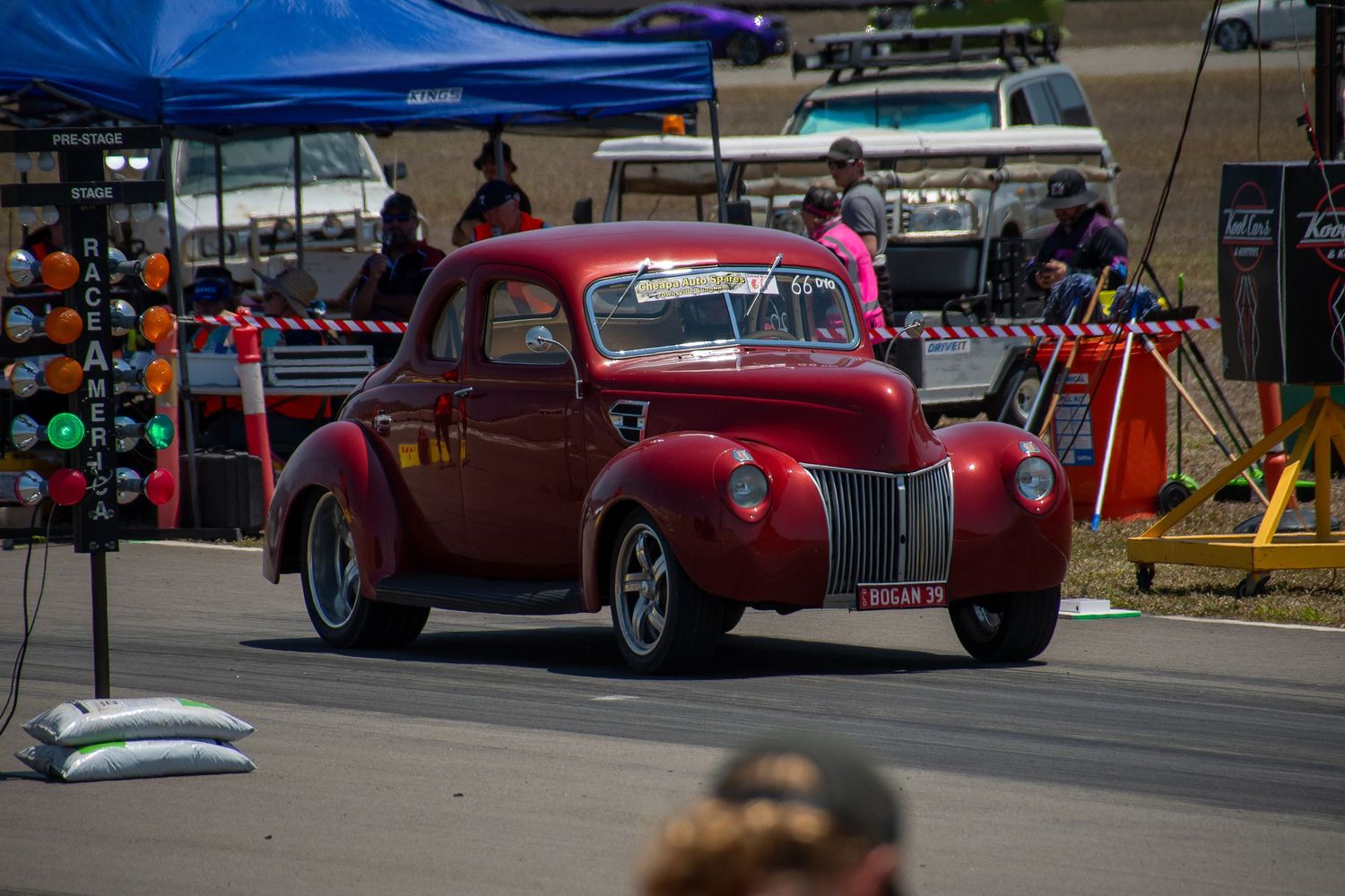 A shiny red vintage hot rod cruises through a street event with colorful displays in the background.