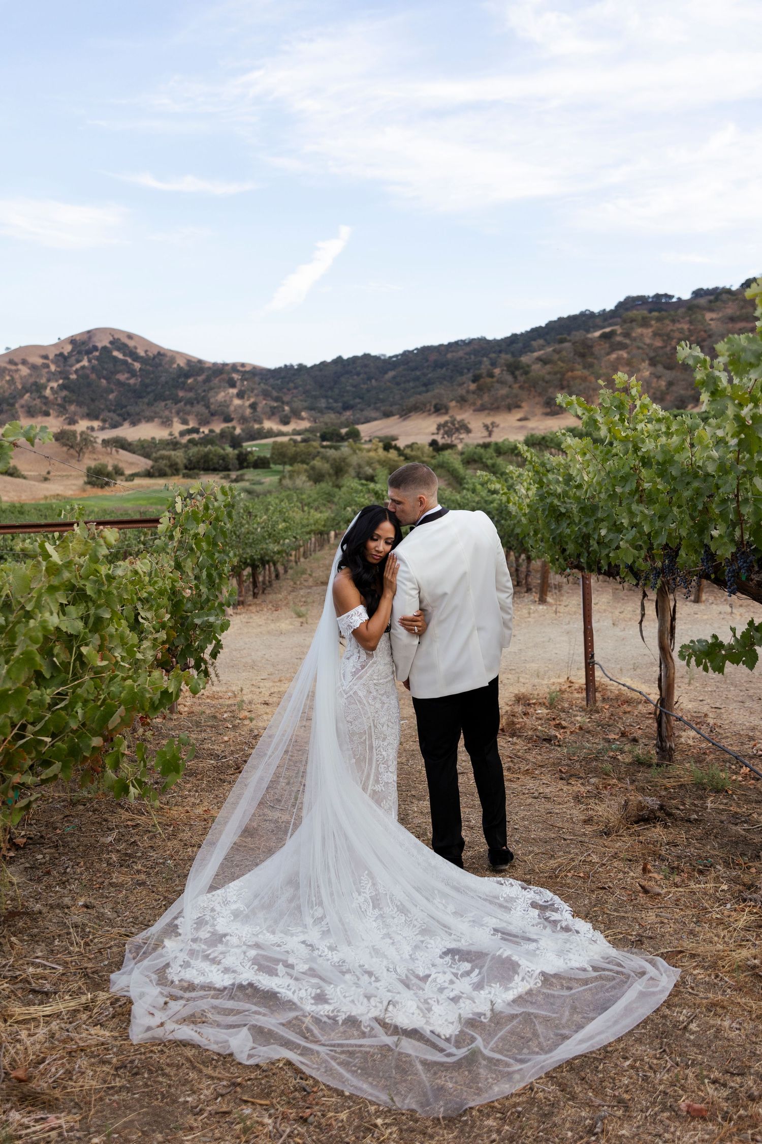 A couple in wedding attire walks through vineyard rows with a flowing lace train trailing behind.