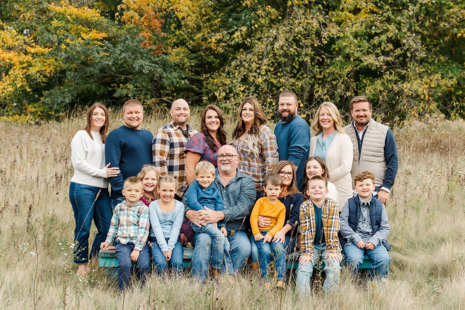 A group of people wearing casual clothing sit together on grass with autumn foliage in the background.