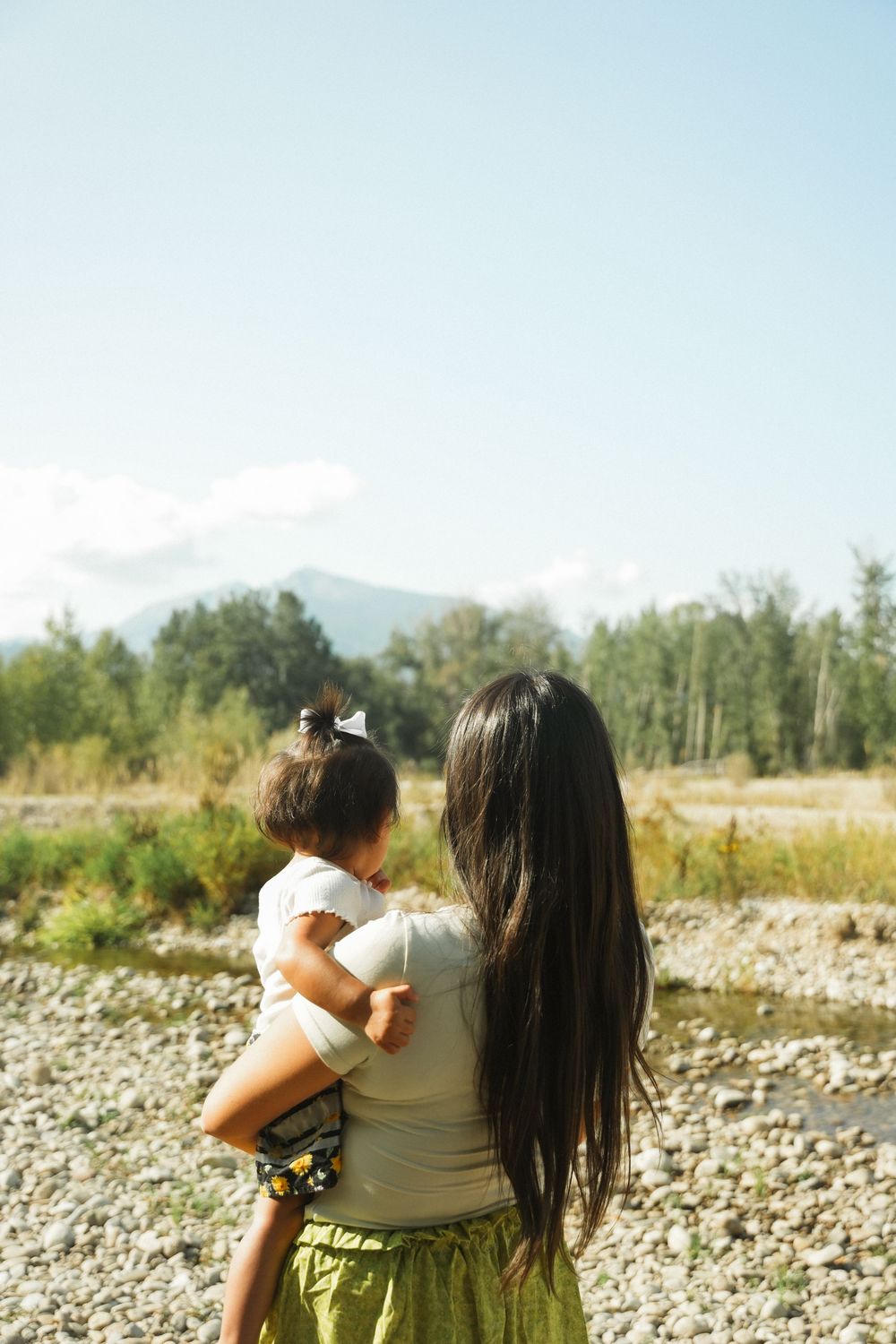 Mother and child walking together on rocky riverbank on sunny day while enjoying outdoor nature.