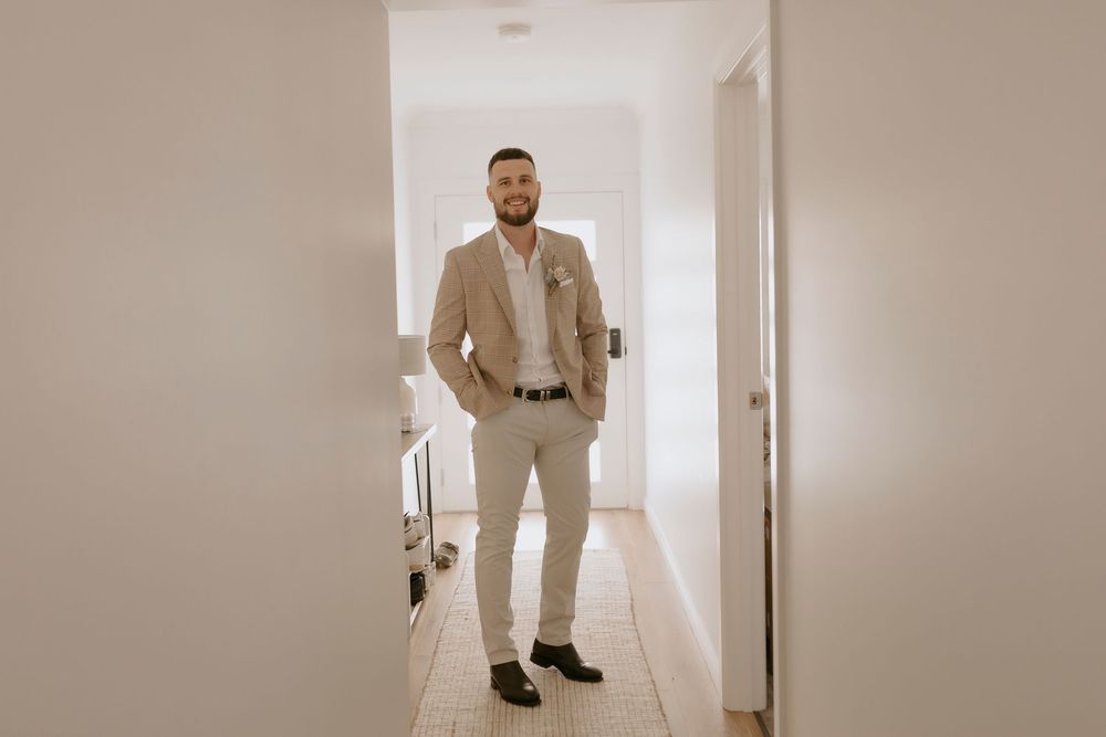 A series of photos showing a groom getting ready in a beige suit and tie in a bright hallway.