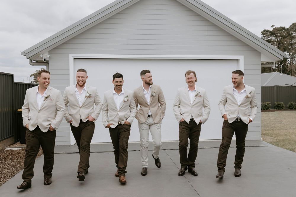 A group in cream-colored jackets and dark pants pose casually in front of a white garage door.